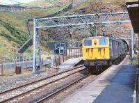 View from the staggered westbound platform at the closed Woodhead Station on 15 August 1979 as 76034+76031 emerge from Woodhead tunnel with a Trans - Pennine coal train.<br><br>[Peter Todd 15/08/1979]