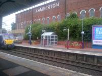 NHS planters on the eastbound platform at Coatbridge Sunnyside on 8 September 2015, with the Lamberton Building in the background. The train arriving is the 0910 Helensburgh Central - Edinburgh Waverley.<br><br>[John Yellowlees 08/09/2015]