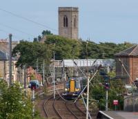 A Ardrossan Harbour bound 380108 approaches Ardrossan South Beach viewed through electrical clutter. The tower of St Cuthbert's Church looks on.<br><br>[Ewan Crawford 10/08/2017]